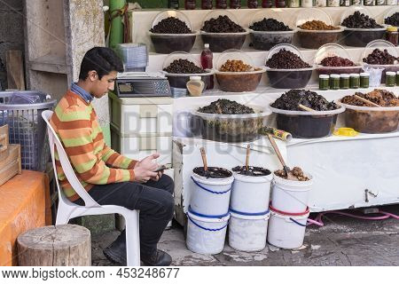 Masuleh, Iran - December 22, 2017 Teenager Seller Assistant Looking His Mobile Phone In Pickles Stor