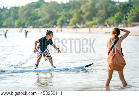 Amature Surfman Surfs The Wave At Kata Beach, Phuket Province Thailand. While A Blur Woman Is Taking