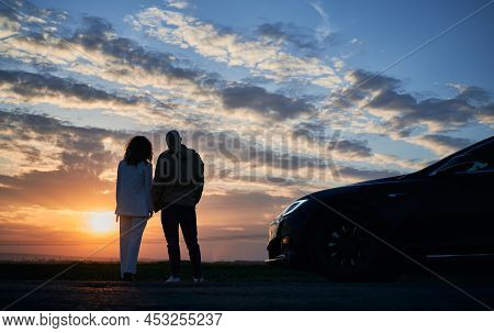 Rear View Of Romantic Couple Who Arrived In Black Car Out Of Town To Admiring Sunset Over Fields At 