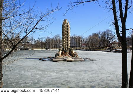 Moscow, Russia - 14 April, 2022: Fountain Golden Spike Ear Snow View On Vdnkh Park Against Blue Sky 