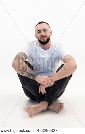 Young Happy Bearded Man With Short Dark Hair In White T-shirt, Black Jeans, Sitting, Bending Knees O