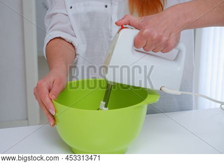 Young Woman Baking A Cake In The Kitchen. Female Using A Handheld Mixer To Whisk The Fresh Ingredien