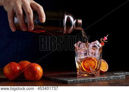 Alcohol Cocktail Pouring From Shaker Into Gless With Orange Peel Ice On Stone Desk.