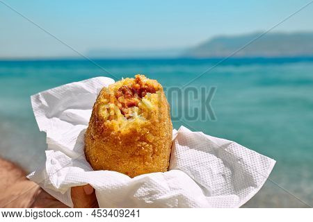 Typical Sicilian Street Food On Seaside Background. Hand Of Man Holding Hot Palatable Arancina (deep