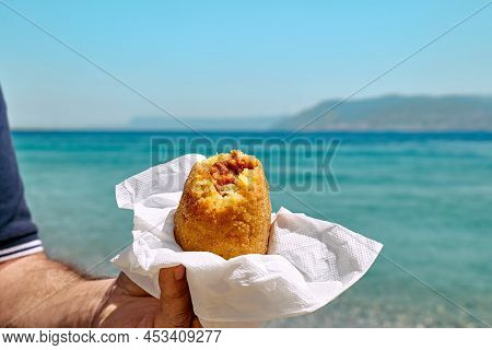 Typical Sicilian Street Food On Seaside Background. Hand Of Man Holding Hot Palatable Arancina (deep