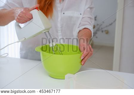 Young Woman Baking A Cake In The Kitchen. Female Using A Handheld Mixer To Whisk The Fresh Ingredien