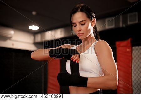 Young Woman Wraps Her Fists In Black Bandages For Kickboxing Gloves Against A Black Boxing Hall