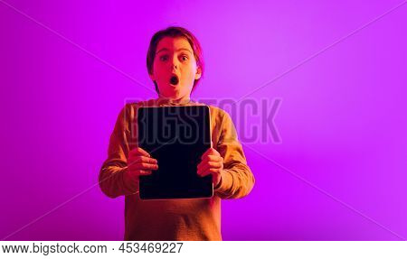 Portrait Of Boy, Child With Expressive Emtions Holding Tablet Isolated Over Pink Background In Neon 