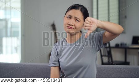 Asian Woman Annoyed Looking At Camera Showing Thumb Down And Sitting On Sofa In Living Room At Home.