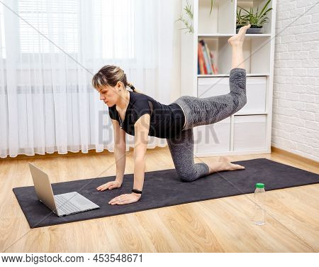 Young Sporty Woman Practicing Yoga. Woman Exercising At Home In Front Of Her Laptop, Stretching Her 