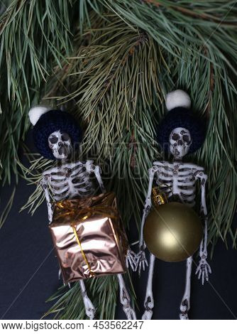 Christmas Concept - Two Funny Skeletons In Skully Hats With Christmas Ball And Gift Against Fir Tree