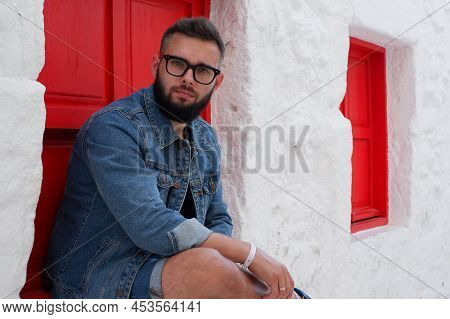 Portrait Of A Handsome, Attractive, Young, Stylish Bearded Man In Glasses And A Denim Jacket Sitting