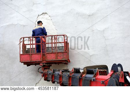 Worker In Hydraulic Lifting Ramp Repair The Building Wall. Builder On Lifting Platform Plasters The 
