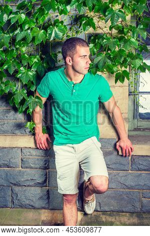 Young Man Waiting. Wearing A Green Short Sleeve Henley Shirt, Light Yellow Shorts, Bending A Leg, A 