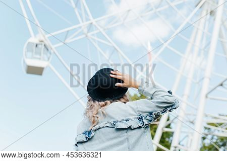 Young Hipster Woman In Hat And Denim Jacket Looking At Ferris Wheel In City Amusement Park On Sunny 