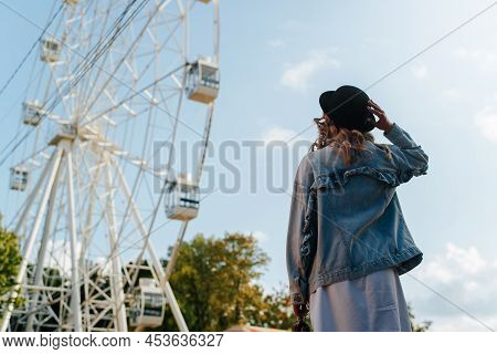 Young Curly Hipster Woman Holding Hat And Looking At Ferris Wheel In City Amusement Park On Sunny Da