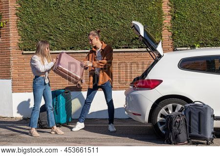 Young Cuple Trying To Fill The White Car Trunk Full Of Suitcases. Girl Passing Boy Luggage On Hands 