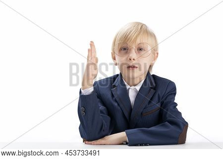 Primary School Student Sits At Table Or Desk With Hand Raised. Portrait Of Straight-a Student Boy Is