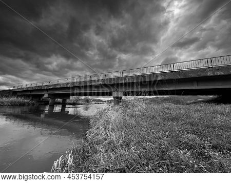 Wide Angle Monochrome Image, Concrete Bridge Over Olt River, Forming Dark Clouds On The Sky.