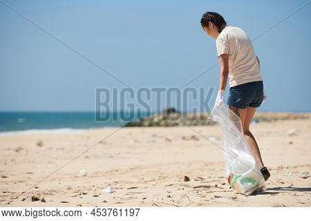 Young Woman Walking On Beach With Big Plastic Bag And Collecting Trash Brought By Sea Wave