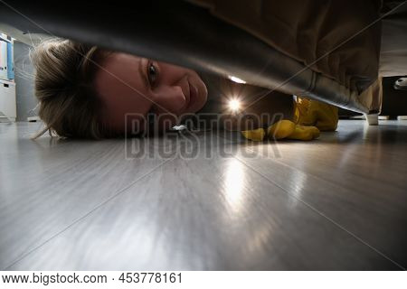 Close-up Of Woman Using Flashlight To Look For Something Under Bed. Female In Protective Gloves Clea