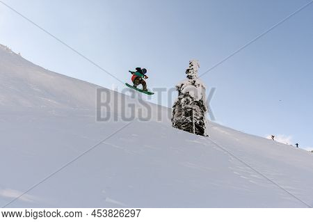 Active Snowboarder In Bright Overalls Slides Down The Mountain On Splitboard And Jumping Up