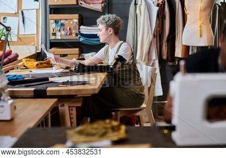 Young Female Fashoin Designer With Partial Arm Looking Through Sketches Of New Clothes While Sitting