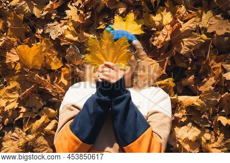 Caucasian Person Laying Down On Autumnal Leaves And Hiding Their Face Behind Big Yellow Leaf. Outdoo