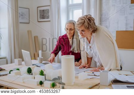 Mature Women Eco Architects With Model Of Modern Bulidings And Blueprints Working Together In Office