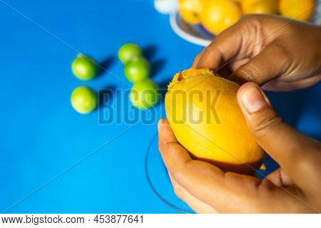 Closeup Of The Hand Of A Latin Woman Peeling A Mango With A Blue Background With Jocotes And Typical