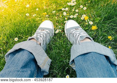Legs In Sneakers And Denim Trousers On Grass With Spring Or Summer Daisies. Ecological Natural Shoes