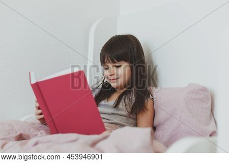 One Todder Girl 5 Years Old Brunette With Long Hair Sits In Bed And Reads A Book And Smile.