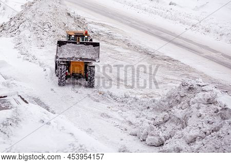 Small Snow Removal Vehicle Removing Snow On City Square. Yellow Or Orange Tractor Cleaning The Snow 
