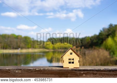 Miniature Wooden Cabin By Against A Beautiful Lake Landscape Background. Lake Cottage Holidays Conce