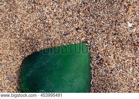 A Piece Of Glass On The Sand. A Shard Of Green Frosted Glass From A Bottle On A Seashell Beach.  A P