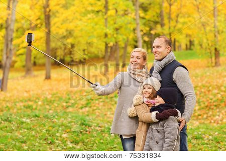 family, childhood, season, technology and people concept - happy family taking selfie with smartphone and monopod in autumn park