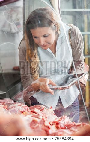 Mature woman holding digital tablet while choosing meat at butchery