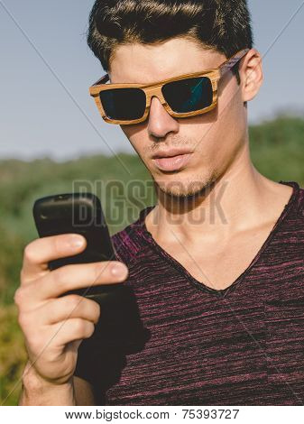 Model Man Portrait With Wooden Sunglasses Outdoors. Man Is Using A Smartphone.