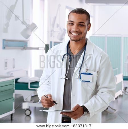 Happy smiling afro american doctor at hospital room with tablet, Looking at camera, standing, wearing stethoscope and lab coat.