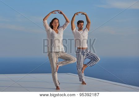 young couple practicing yoga at sunset in modern home terace with ocean and sunset in background