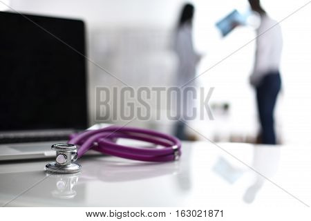 Laptop and stethoscope on the desk doctors standing in the background.