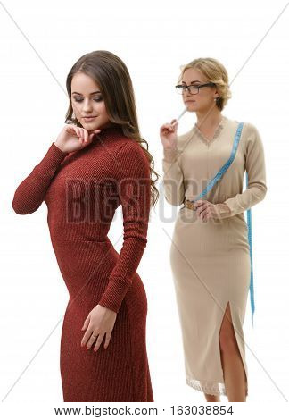 Young sexy dressmaker in slinky beige dress takes measurements of her cute slim long haired client in sexual knitted winy gown studio shot