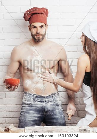 Pretty girl or beautiful woman cute helper sprinkles flour on handsome man or muscular chef cook baker with sexy muscle torso body with six packs and abs on kitchen wall