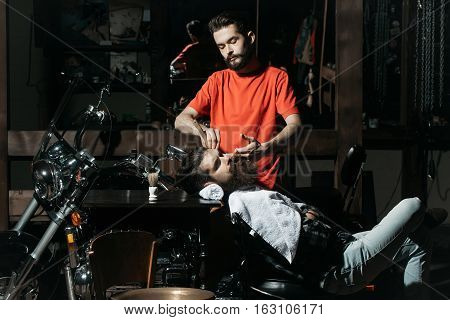 Handsome barber or shaver shaves beard to bearded man with vintage razor with open blade. Male customer sits in chair in dark garage with motor bike