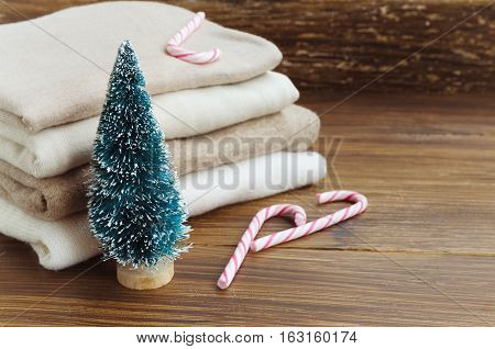 Stack of knitted clothes in beige tones. Cashmere sweaters wool cardigans little Christmas tree and candy cane on a wooden background. Concept of Winter time and Christmas holiday. Copy space.