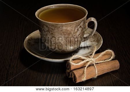 Brown tea cup and saucer, a bundle of cinnamon and sugar on dark wooden background.