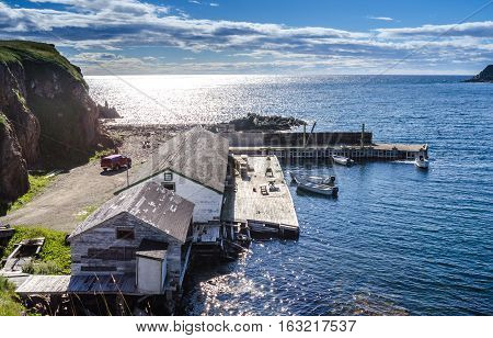 Twillingate, Newfoundland, man works on the engine of one of 4 boats tied up to a wooden, cliff-side dock house for the day, bright sunshine on calm coastal water.