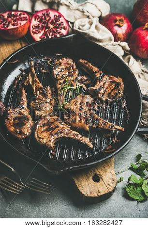 Barbecue dinner. Grilled lamb meat chops with onion and rosemary in cast iron pan served with fresh pomegranates and mint on wooden board over grey background, selective focus. Slow food concept