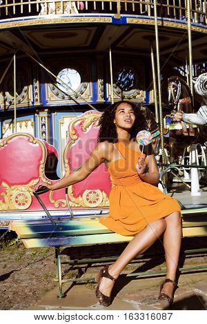 cool real african american teenage girl with candy near carousels at amusement park, lifestyle people concept close up