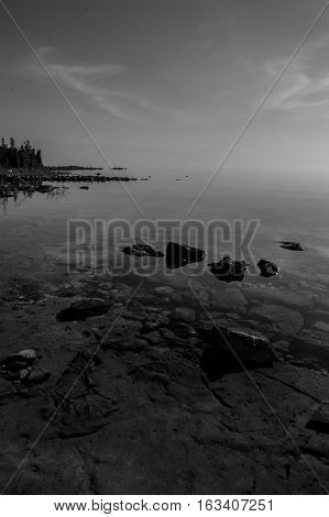 Dead calm afternoon image of clear lake huron water and limestone rocks along shoreline. Clouds reflecting in a zign zag. b/w
